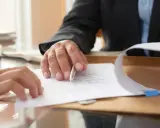 Dwie osoby przy biurku omawiają dokumenty. Jedna z nich, w garniturze, wskazuje długopisem na tekst. To może być poświadczenie notarialne przy wynajmie mieszkania.