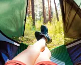 Nogi w butach trekkingowych odpoczywają w namiocie. Słońce przebija się przez drzewa. Idealne miejsce, by zanocuj w lesie.