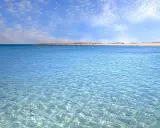 Crystal clear turquoise water with gentle ripples, leading to a sandy beach and distant dunes under a blue sky with scattered clouds.