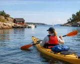 En person i gul kajak njuter av att **padla kajak** i en vacker skärgårdsmiljö med röda hus och segelbåt i bakgrunden.