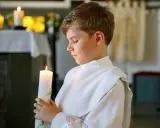 Chłopiec w białej albie trzyma zapaloną świecę podczas uroczystości komunii świętej.