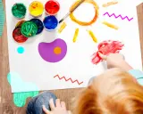Bambino dipinge un sole con le dita, circondato da vasetti di colori. Un esempio di giochi popolari per bambini che stimolano la creatività.