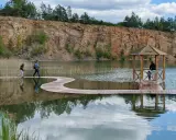 Malownicza trasa Gródka Jaworzno z drewnianym molo i altanką. Dwie osoby spacerują po falistej kładce nad wodą.