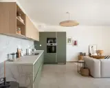 Cuisine moderne avec des armoires vert olive, un plan de travail en marbre et des étagères en bois clair. Un salon adjacent est visible.