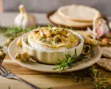 Pieczony camembert w naczyniu, ozdobiony ziołami i orzechami, z czosnkiem i pieczywem w tle.