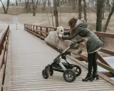 Kobieta na drewnianym moście poprawia gondolę wózka. Zastanawiasz się, jaki wózek wybrać mamaginekolog? Ten model wygląda na solidny.