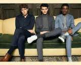 Three men in suits sit on a couch. The man on the left wears dark dress pants, while the other two wear suit pants.