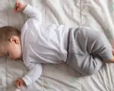 Un neonato non dorme, ma giace sdraiato su una coperta con un pigiamino bianco e grigio.