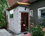 Rozbudowa domu na zgłoszenie w toku. Nowa ściana z bloczków, drzwi i okno czekają na wykończenie. Wokół kwitną kwiaty.