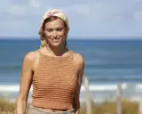 Woman in a crocheted top and headscarf smiles at the camera on a beach.