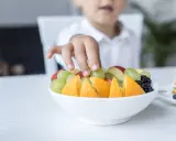 Małe rączki sięgają po miskę pełną owoców: pomarańczy, truskawek, jabłek i winogron dla niemowlaka.