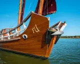 Ein hölzernes Dschunke Schiff gleitet auf ruhigem Wasser unter blauem Himmel. Die Segel sind teilweise gesetzt.