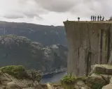 Grupa ludzi na krawędzi klifu w **Stavanger, Norwegia**, z widokiem na fiord i góry.