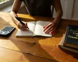 Une personne écrit dans un carnet, une calculatrice à côté. Un livre "The Complete Guide to Numerology" est posé sur la table.