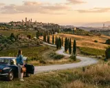 Donna con mappa accanto a un'auto d'epoca in un paesaggio collinare toscano, con borghi medievali e vigneti. Un perfetto itinerario toscana.