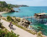 Ciclista lungo la via dei trabocchi, con un trabocco in legno sull'acqua cristallina e una spiaggia rocciosa.