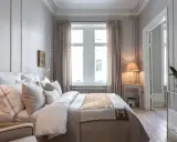 Ett elegant sovrum med en stor säng bäddad med vita lakan och en brun pläd med H-mönster. Vackra möbler till sovrummet som ett litet sidobord med lampa och en orkidé.