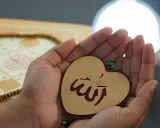 Hände halten ein Herz mit dem Wort "Allah" in arabischer Schrift, ein Symbol für ikhlas sure. Gebetskette und ein Buch liegen im Hintergrund.