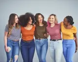 Cinque amiche sorridono, vestite con jeans e magliette colorate, tra cui spiccano tonalità marrone e bianco.