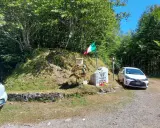 Monumento commemorativo con bandiera italiana e corona d'alloro al passo delle radici, circondato da alberi e con un'auto parcheggiata.