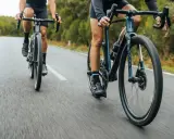 Dwie osoby na rowerach szosowych ROMDO RATT pędzą po asfaltowej drodze wśród zieleni.