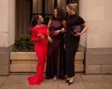 Three women in elegant gowns, discussing what not to wear to a wedding. One in red, one in plum, and one in black.