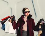 Donna con occhiali da sole e rossetto rosso, avvolta in un cappotto bordeaux, porta borse della spesa, simbolo di moda veloce.