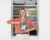 Kobieta z książką "The Folding Lady" pokazuje, jak składać koszulki, stojąc przed otwartą szafą pełną schludnie ułożonych ubrań.