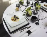 Une table de restaurant dressée avec élégance, un plat de poisson en vedette, des verres noirs et des fleurs.