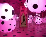 A woman walks through a mirrored room filled with pink polka-dotted spheres, a signature of Yayoi Kusama's immersive art.