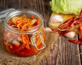 Słoik z kimchi na tle desek, obok kapusta, czosnek i papryczki chili.