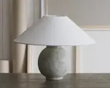 Bordslampa med rund, texturerad fot av lufttorkande lera, målad i grågröna toner. En vit lampskärm sitter ovanpå.