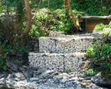 Estructura de muro de gavion con piedras blancas, rodeada de vegetación y rocas junto a un río.