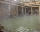 Dampf steigt aus dem Becken der römischen Therme auf. Säulen und Bögen umgeben das Wasser, in dem Enten schwimmen.