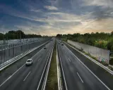 Szeroka autostrada z samochodami w oddali, otoczona zielenią i barierami dźwiękochłonnymi. Jazda autostradą pod pochmurnym niebem.