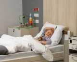 Uśmiechnięte dziecko w szpitalnym łóżku, przytulające misia. Obok zabawka edukacyjna i szafka.