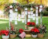Decoración ceremonia civil con un arco de ramas y hojas, colgando tarjetas de invitados. Flores rojas y cestas adornan la base.