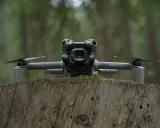 Szary dron DJI Mini 5 Pro z kamerą na gimbalu spoczywa na pniu drzewa w lesie.