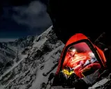 Nocny obóz na zboczu himalajskiego parbatu. W pomarańczowym namiocie rozświetlonym czołówką odpoczywa alpinista, otoczony surowym, ośnieżonym krajobrazem.