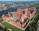 Potężny zamek krzyżacki, największy zamek w Europie, wznosi się nad rzeką Nogat. Czerwona cegła i liczne wieże tworzą imponujący widok.