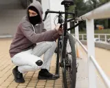 Mężczyzna w masce i kapturze próbuje ukraść rower, odpinając go od poręczy.
