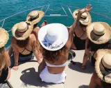 Grupo de amigas en un barco celebrando unas divertidas ideas despedida de soltera. Llevan sombreros de paja con nombres bordados, uno dice "La Novia".