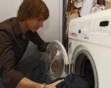 Junger Mann lädt Wäsche in die Waschmaschine. Bald werden die Sitzkissen frisch gewaschen sein.