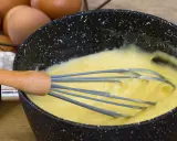 Un fouet mélange une crème pâtissière traditionnelle onctueuse dans une casserole. Des œufs sont visibles en arrière-plan.