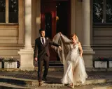 Para młoda podczas sesji zdjęciowej ślubnej schodzi po schodach przed eleganckim budynkiem. Panna młoda w zwiewnej sukni unosi welon, pan młody w garniturze patrzy na nią z uśmiechem.
