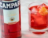 Butelka Campari i szklanka z czerwonym drinkiem, lodem i skórką pomarańczy. Campari to gorzki aperitif.