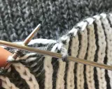 Närbild på patentstickning på rundsticka med grått och vitt garn. En tumme håller i stickan.