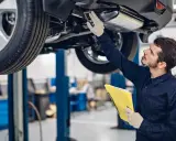 Mechanik w rękawiczkach sprawdza podwozie auta na podnośniku, trzymając żółty segregator. To kluczowy etap, jak sprawdzić auto przed zakupem.