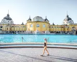 Frau mit Hut spaziert am Beckenrand des Budapester Thermalbads. Das beeindruckende gelbe Gebäude mit Kuppeln bildet die Kulisse.