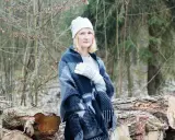 Kobieta w czapce i rękawiczkach z wzorem, otulona grubym pledem. W tle las i ścięte drzewa. Ciepła wełna a bawełna w zimowej stylizacji.